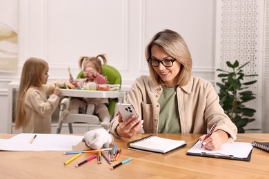 Photo of Work at home. Smiling single mother having videochat by smartphone while her children playing indoors, selective focus