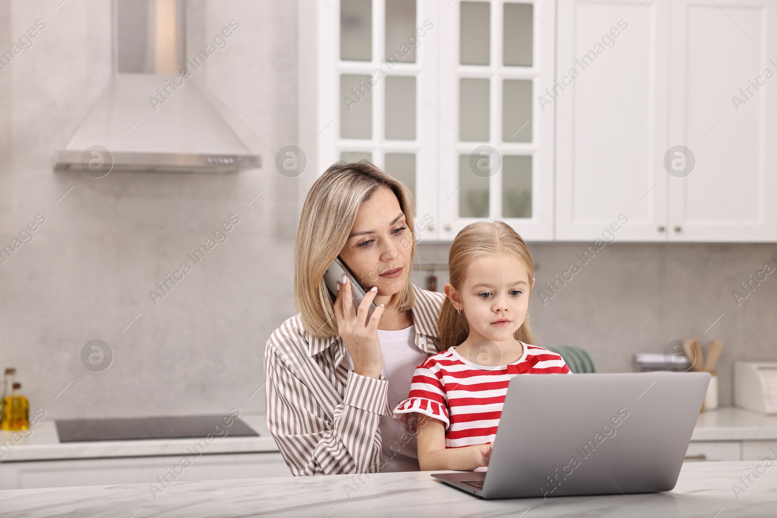 Photo of Work-family balance. Single mother talking by smartphone and her daughter at table with laptop in kitchen