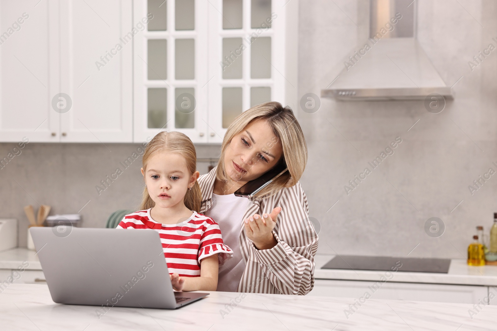 Photo of Work-family balance. Single mother talking by smartphone and her daughter at table with laptop in kitchen