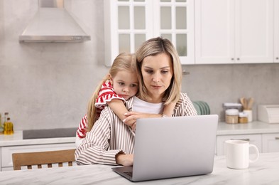 Photo of Work-family balance. Single mother working with laptop and her daughter at table in kitchen