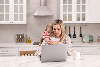 Photo of Work-family balance. Single mother working with laptop and her daughter at table in kitchen