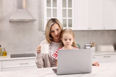 Photo of Work-family balance. Single mother working with laptop and her daughter at table in kitchen