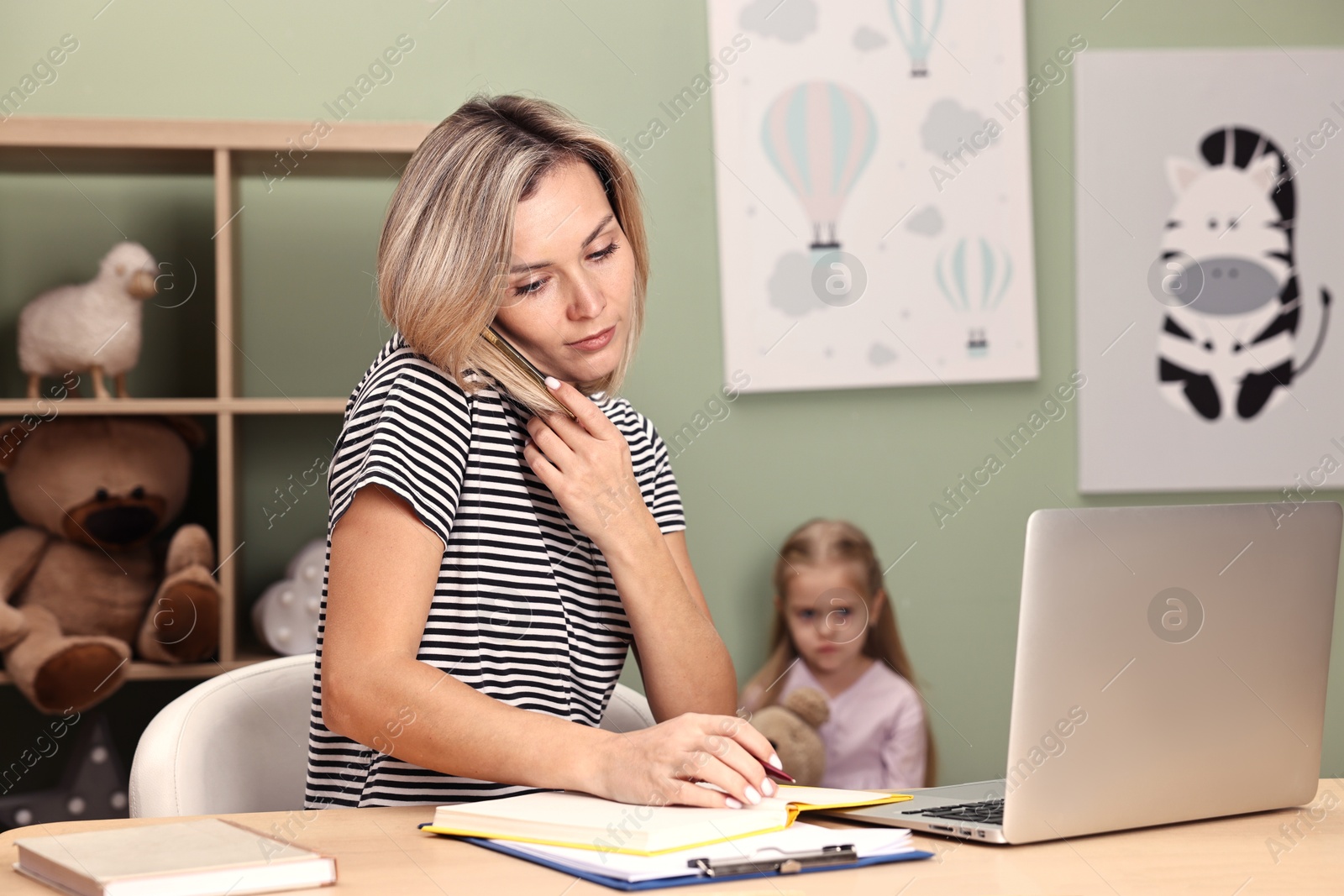 Photo of Work-family balance. Single mother talking by smartphone at table and her offended daughter in child's room, selective focus