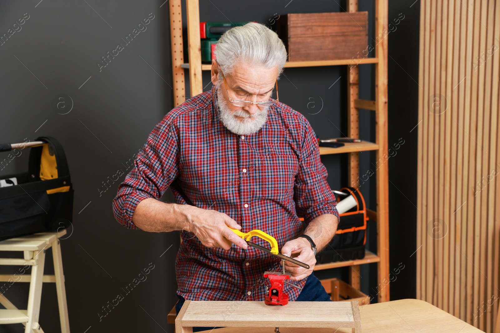 Photo of Relaxing hobby. Senior man with hacksaw and vise assembling wooden shelf in workshop