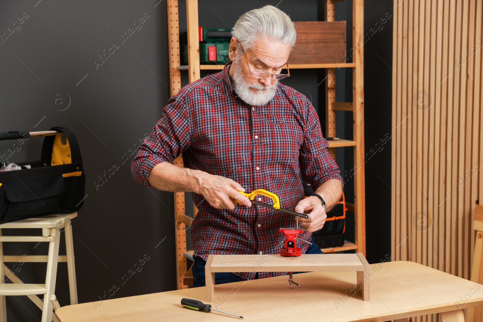 Photo of Relaxing hobby. Senior man with hacksaw and vise assembling wooden shelf in workshop