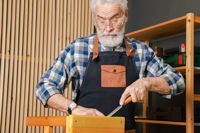 Photo of Relaxing hobby. Senior man repairing wooden stool with chisel in workshop