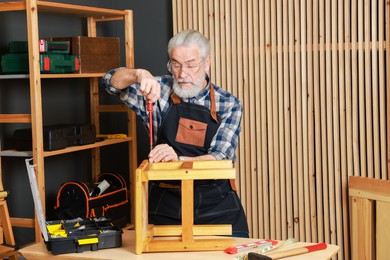 Photo of Relaxing hobby. Senior man repairing wooden stool with screwdriver in workshop