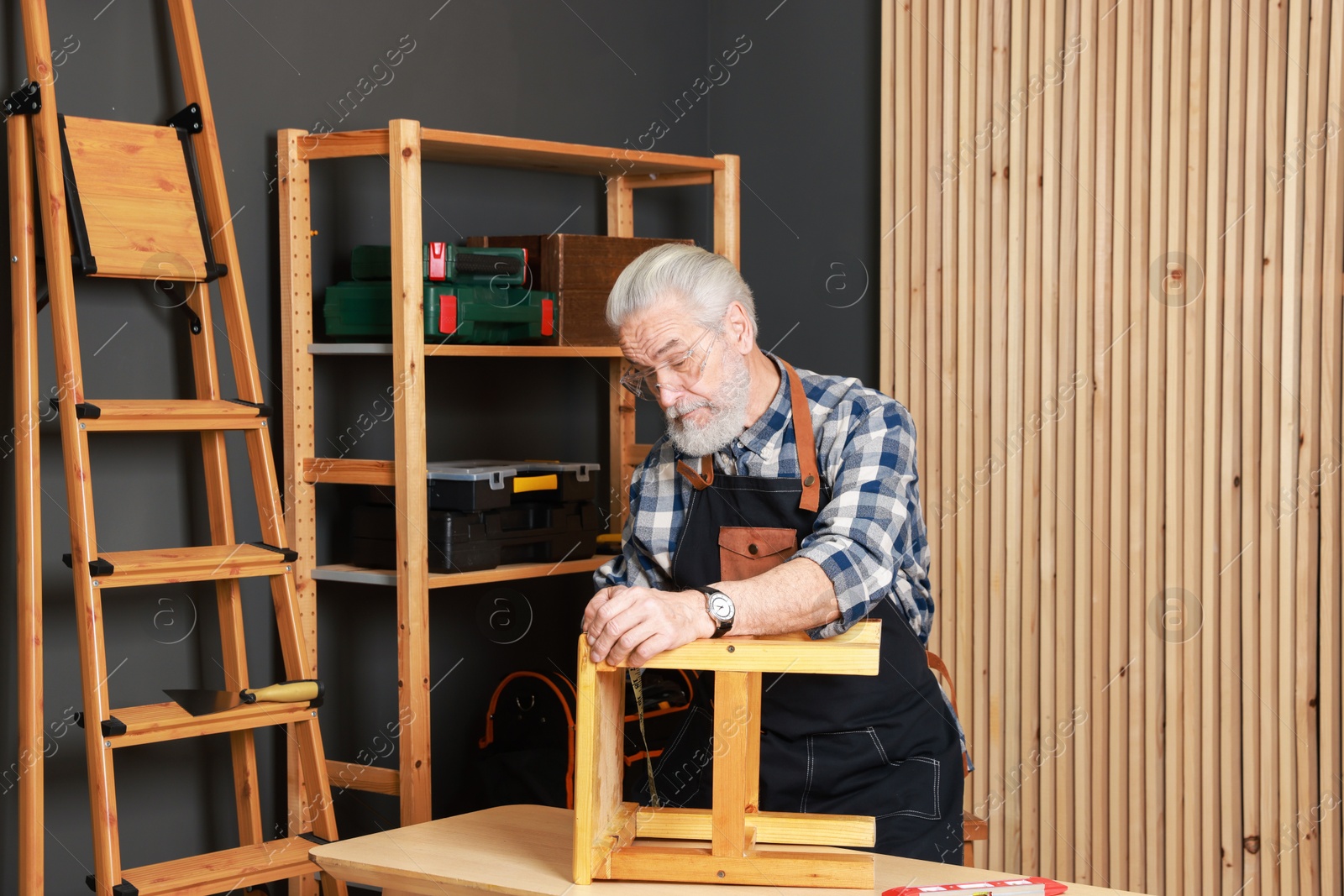 Photo of Relaxing hobby. Senior man measuring wooden stool with tape in workshop