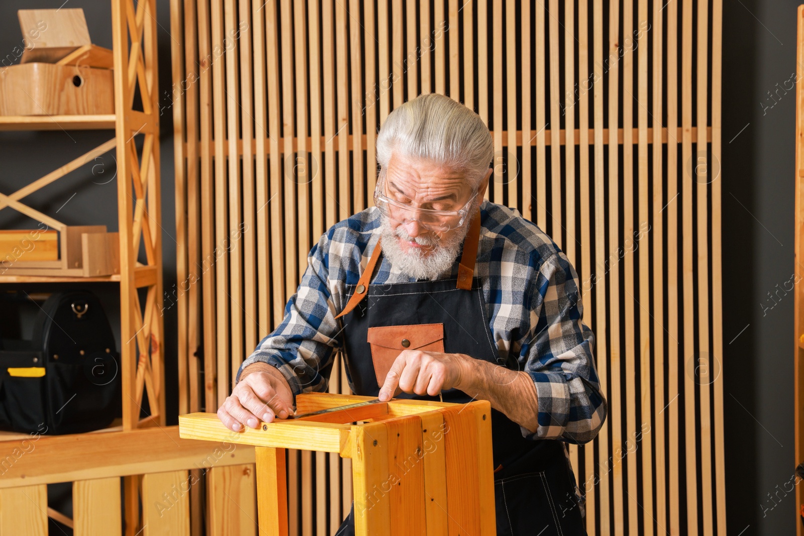 Photo of Relaxing hobby. Senior man measuring wooden stool with tape in workshop