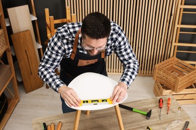 Photo of Relaxing hobby. Man using level tool while repairing stool at wooden table in workshop