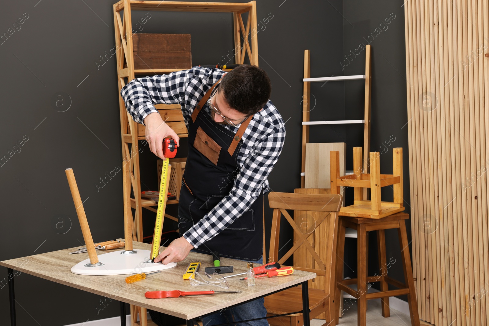 Photo of Relaxing hobby. Man measuring stool with tape at wooden table in workshop