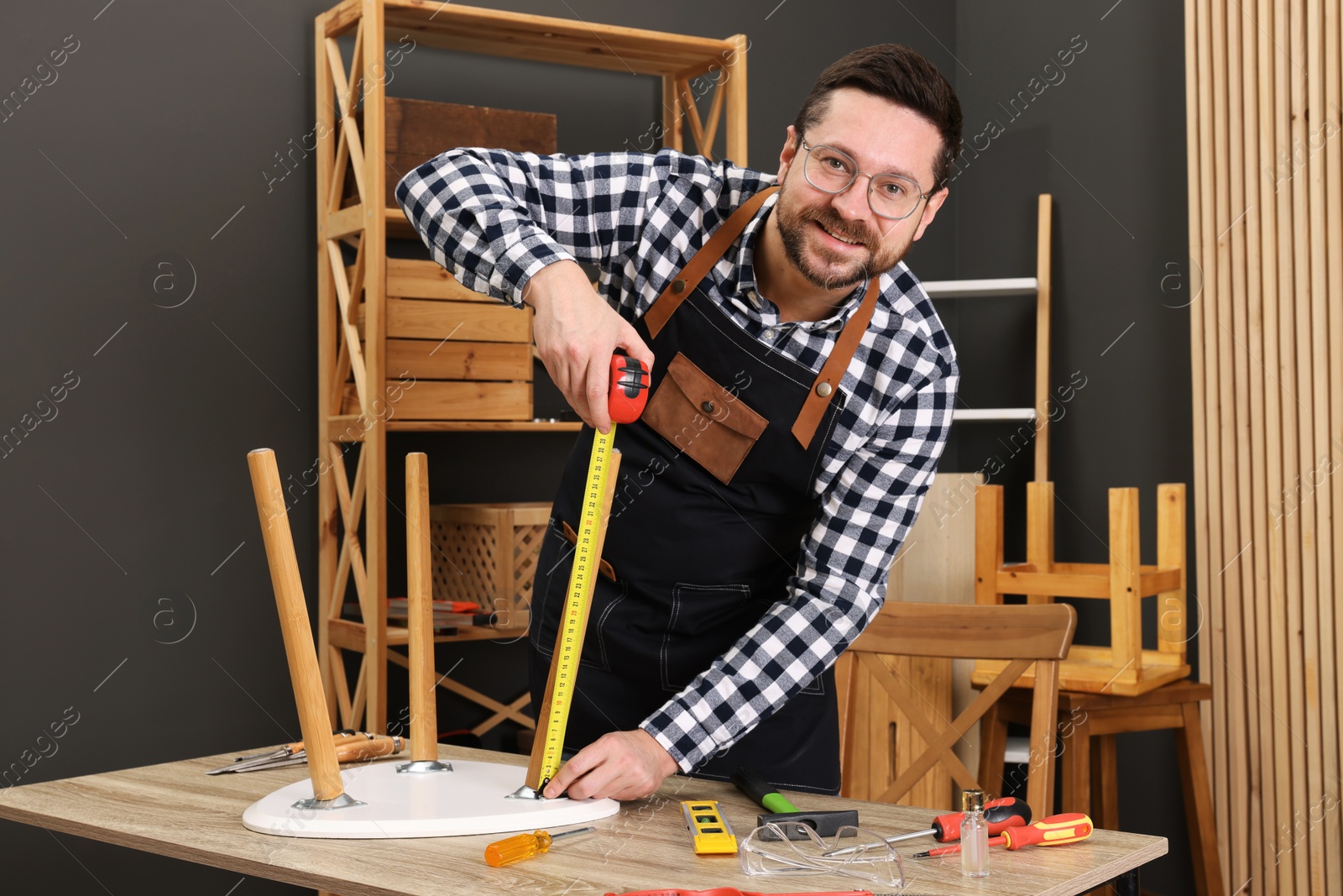 Photo of Relaxing hobby. Man measuring stool with tape at wooden table in workshop