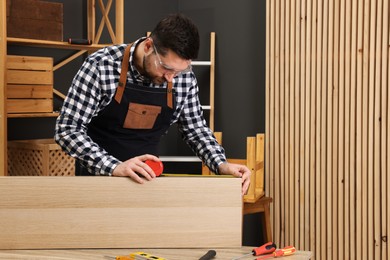 Photo of Relaxing hobby. Man measuring wooden plank with tape at table in workshop. Space for text