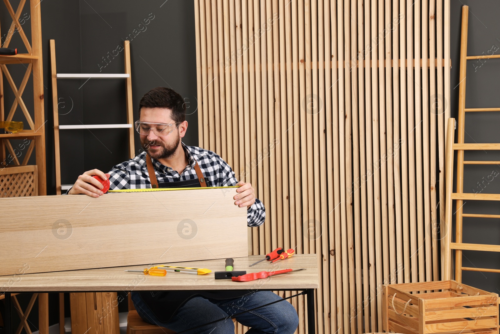 Photo of Relaxing hobby. Man measuring wooden plank with tape at table in workshop. Space for text