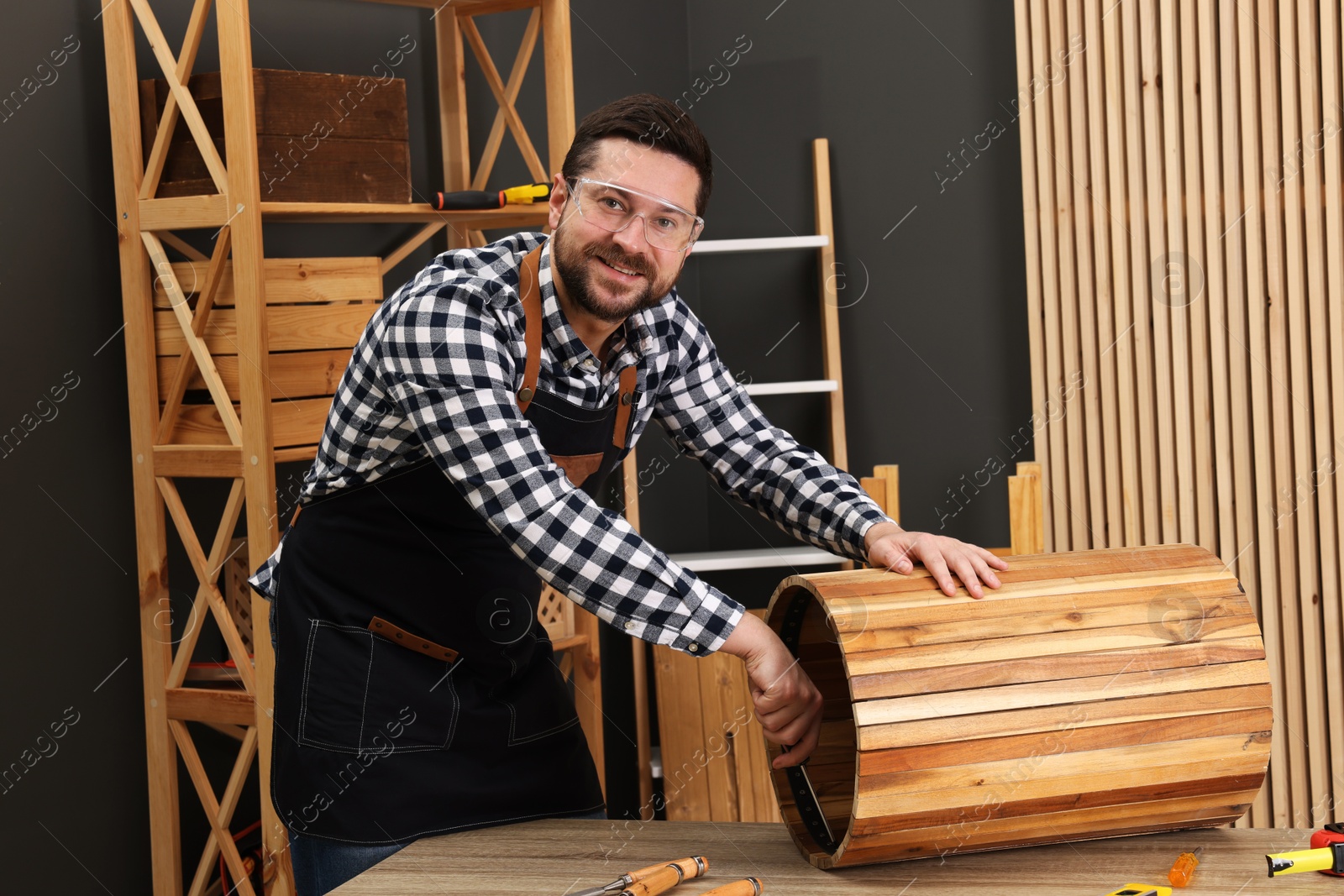 Photo of Relaxing hobby. Smiling man assembling wooden trash can with screwdriver at table in workshop