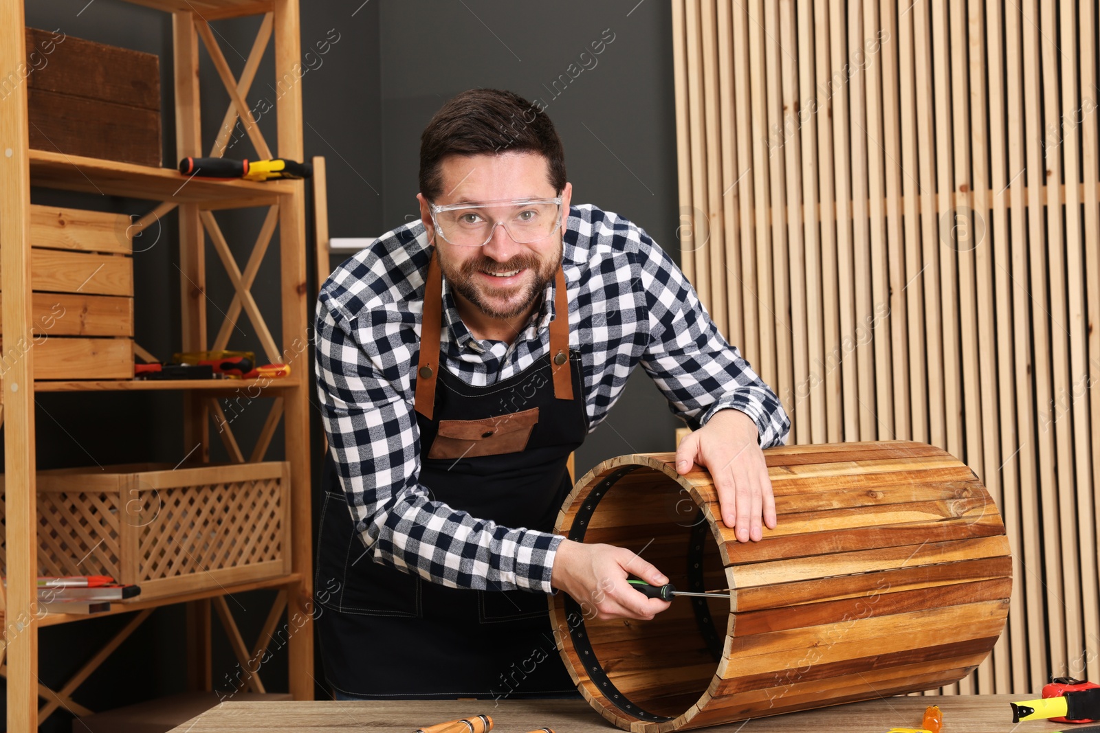 Photo of Relaxing hobby. Smiling man assembling wooden trash can with screwdriver at table in workshop