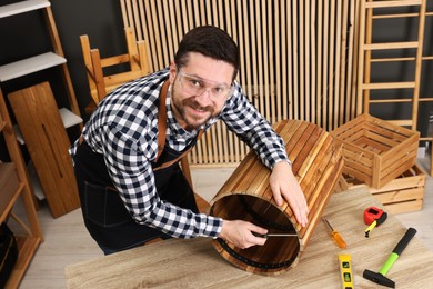 Photo of Relaxing hobby. Smiling man assembling wooden trash can with screwdriver at table in workshop