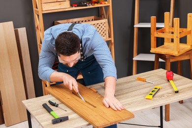 Photo of Relaxing hobby. Man working with wooden plank and chisel at table in workshop