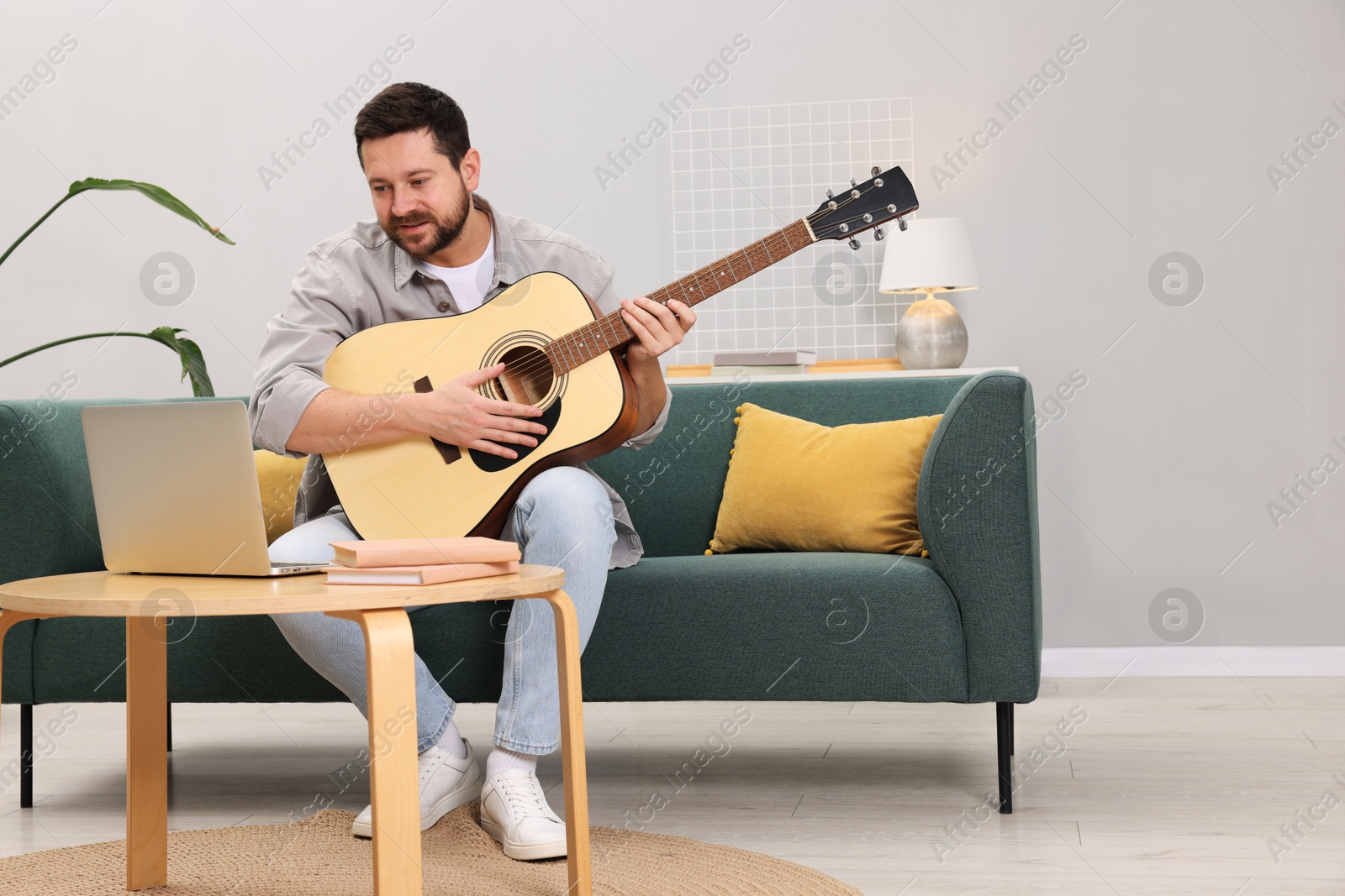 Photo of Relaxing hobby. Man playing guitar with online tutorial by laptop on sofa at home