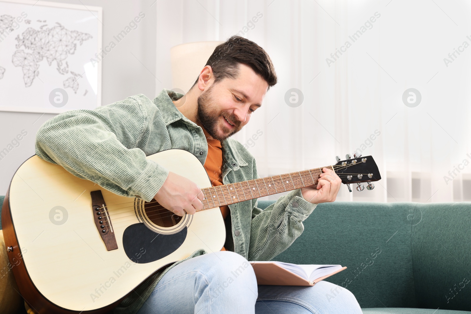 Photo of Relaxing hobby. Smiling man with guitar and book on sofa at home