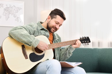 Photo of Relaxing hobby. Smiling man with guitar and book on sofa at home