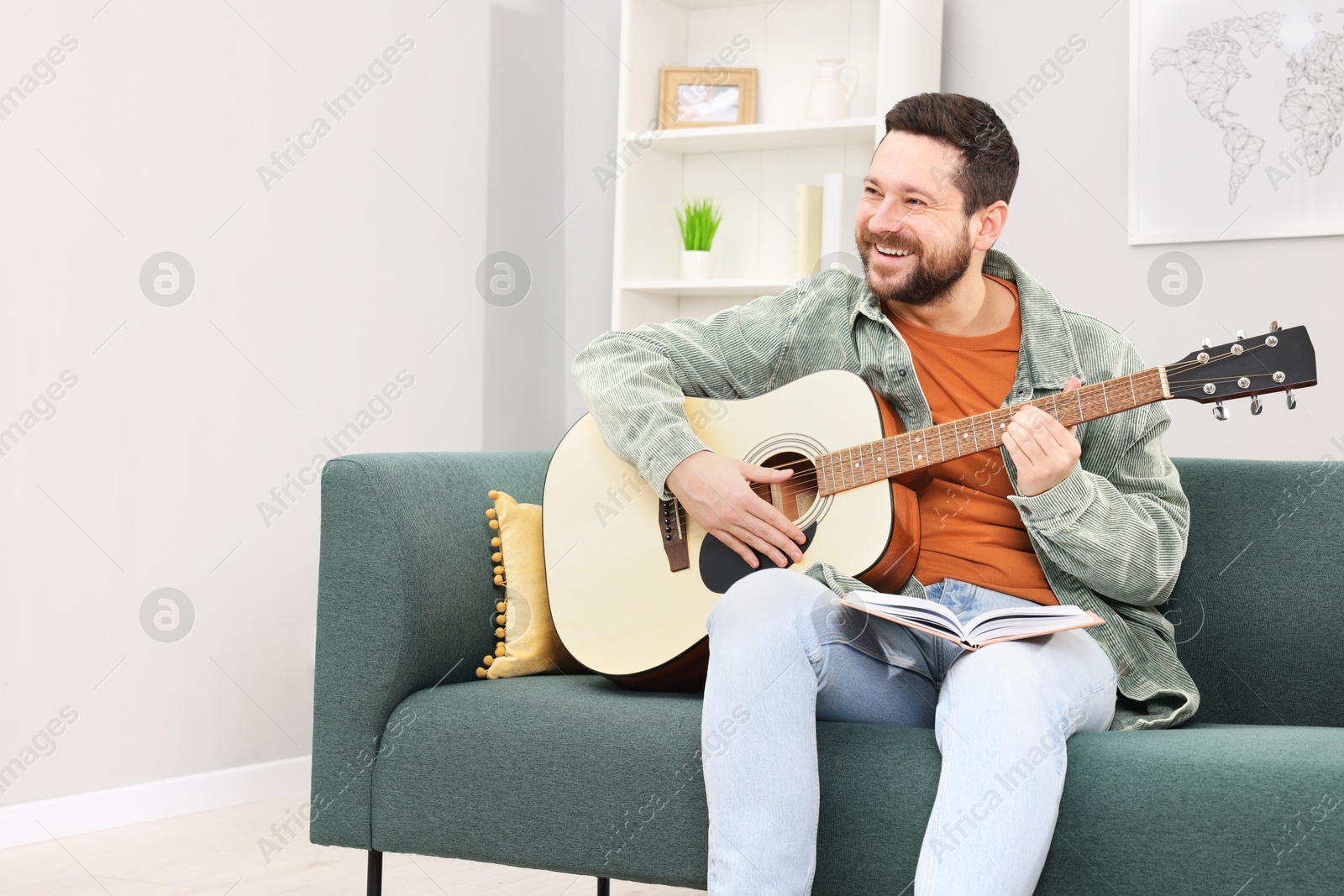 Photo of Relaxing hobby. Smiling man with guitar and book on sofa at home, space for text