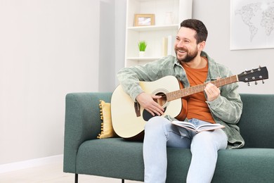 Photo of Relaxing hobby. Smiling man with guitar and book on sofa at home, space for text