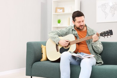 Photo of Relaxing hobby. Man with guitar and book on sofa at home, space for text