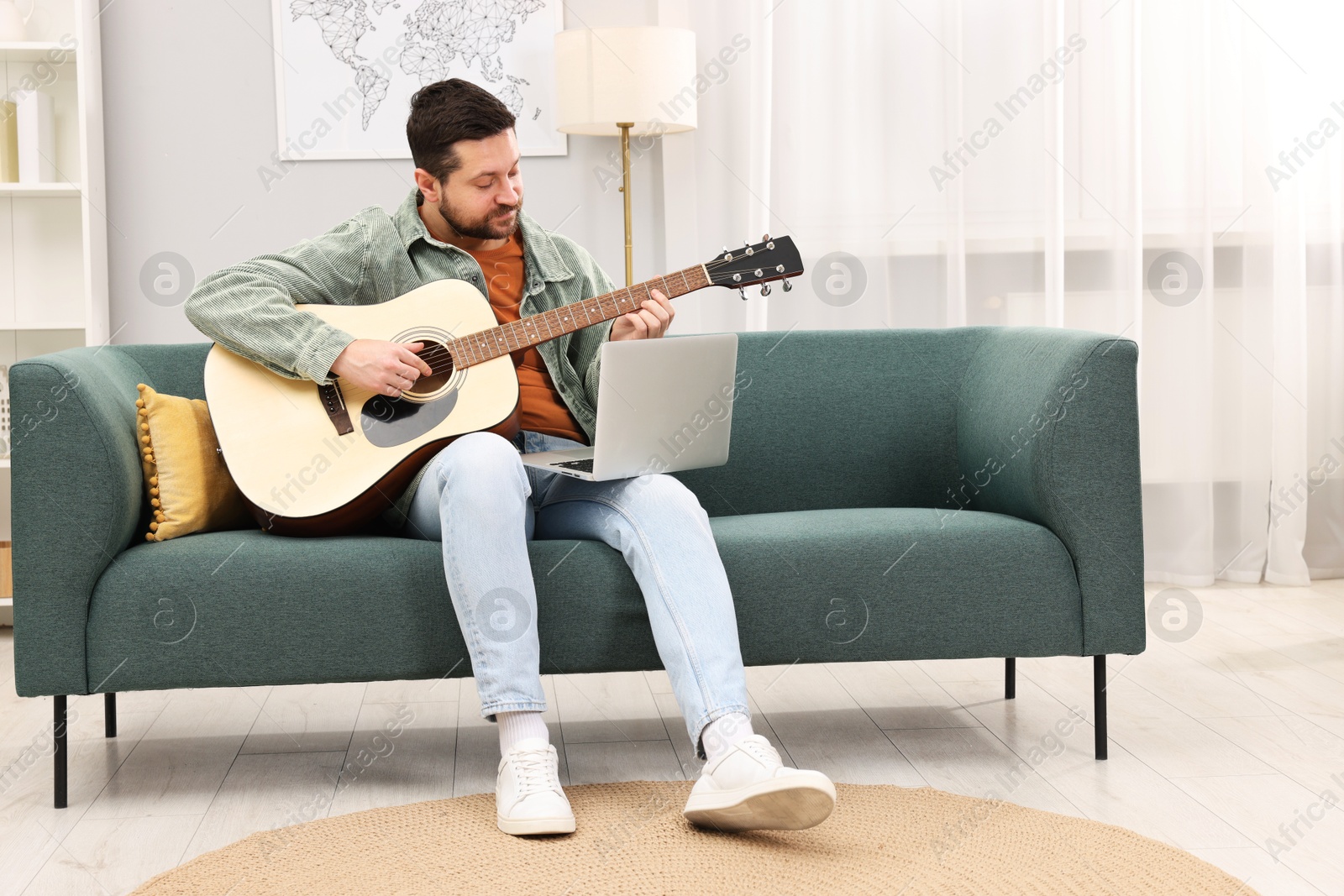 Photo of Relaxing hobby. Man playing guitar with online tutorial by laptop on sofa at home