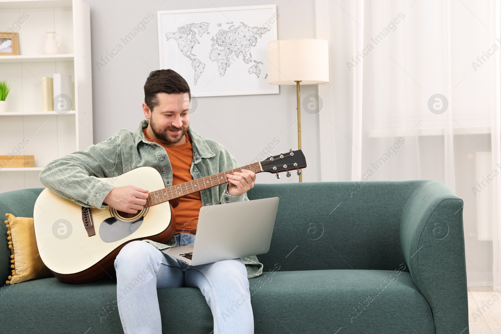 Photo of Relaxing hobby. Man playing guitar with online tutorial by laptop on sofa at home