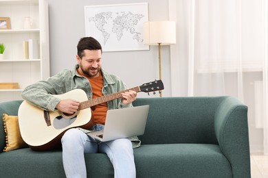 Photo of Relaxing hobby. Man playing guitar with online tutorial by laptop on sofa at home