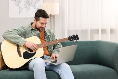 Photo of Relaxing hobby. Man playing guitar with online tutorial by laptop on sofa at home