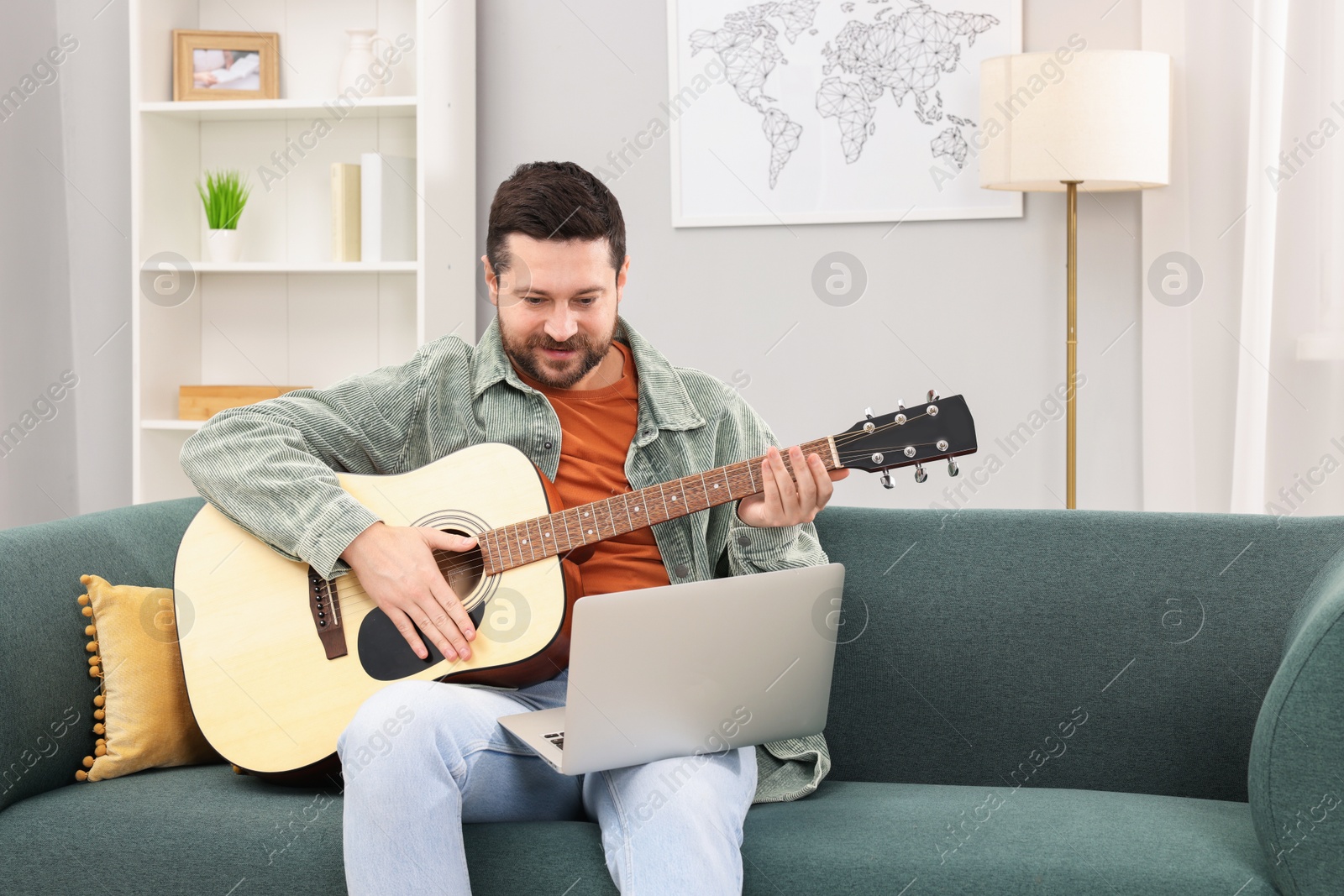 Photo of Relaxing hobby. Man playing guitar with online tutorial by laptop on sofa at home