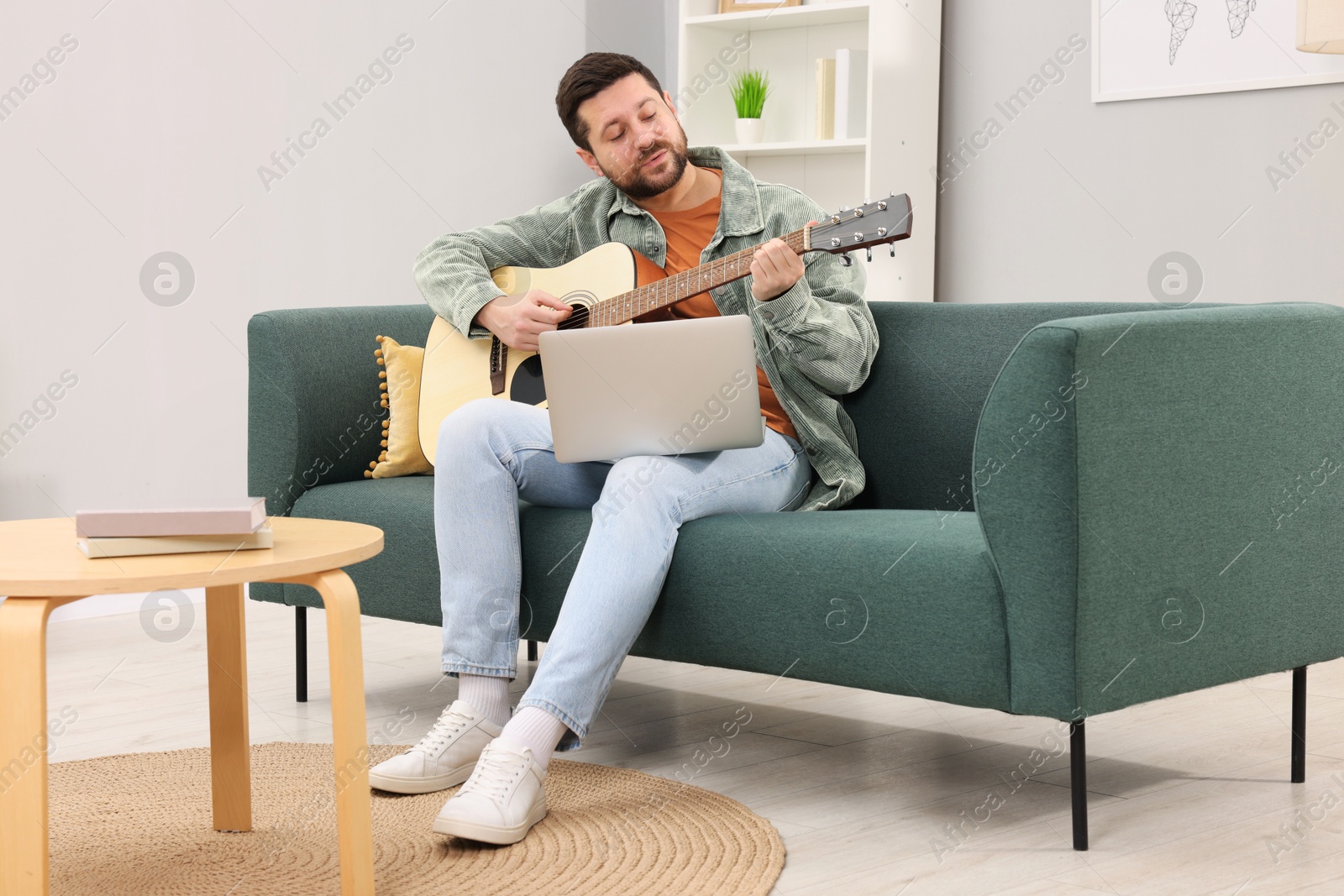 Photo of Relaxing hobby. Man playing guitar with online tutorial by laptop on sofa at home