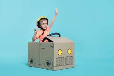 Photo of Little boy showing V-sign while driving car made of cardboard on light blue background