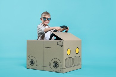 Photo of Little boy in sunglasses driving car made of cardboard on light blue background