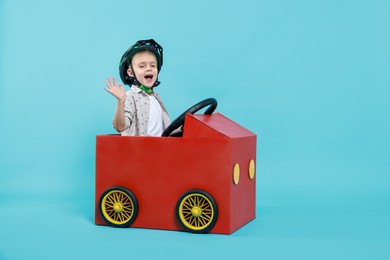 Photo of Little boy waving while driving car made of cardboard on light blue background