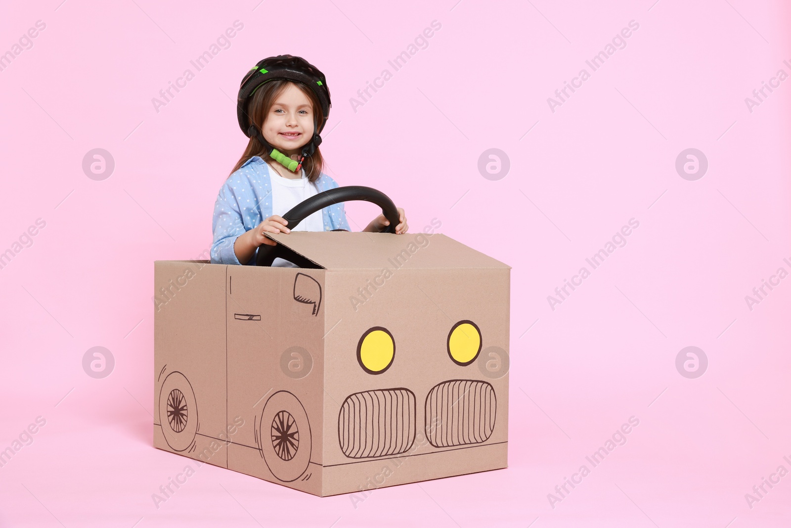 Photo of Little girl driving car made with cardboard on light pink background. Space for text