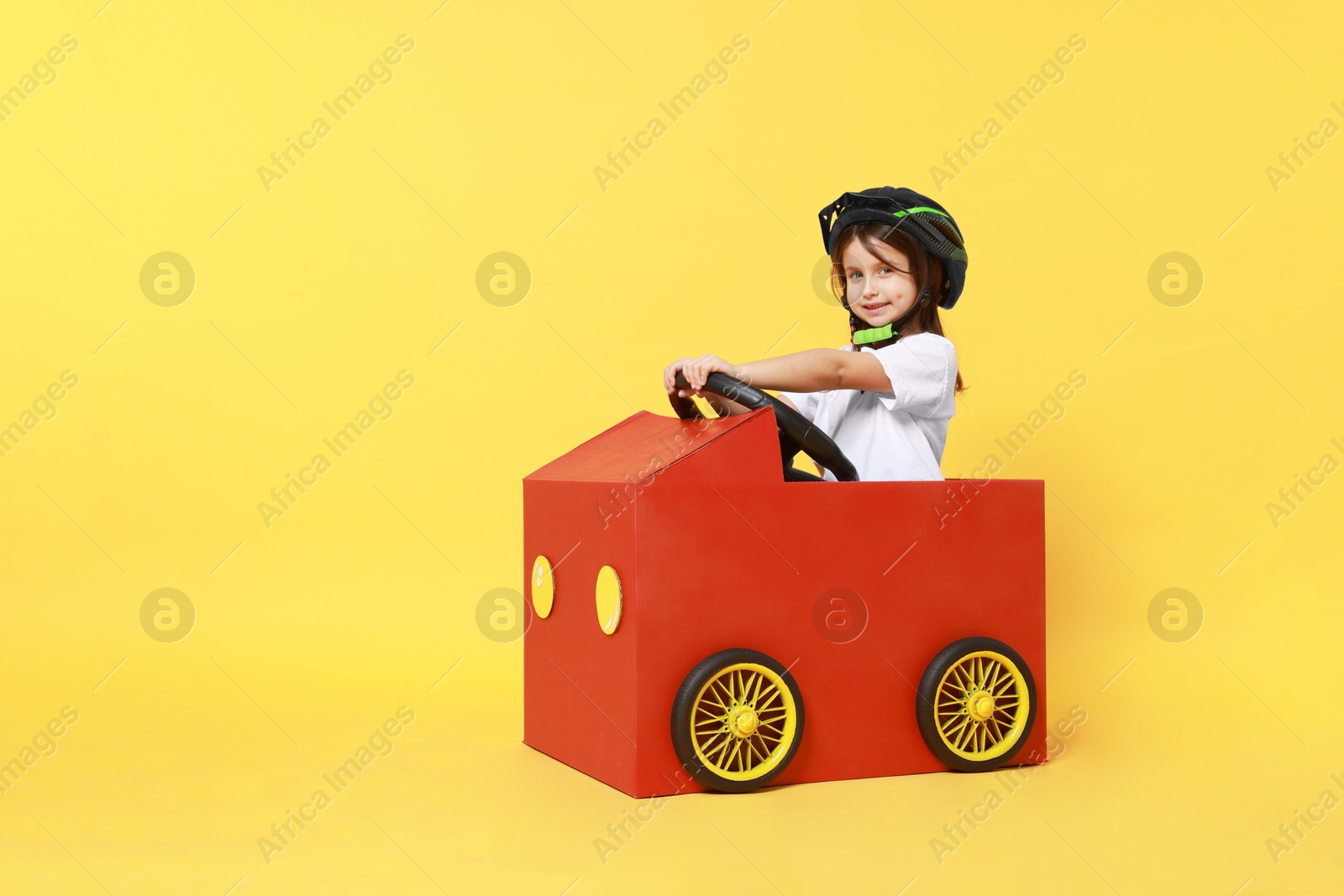 Photo of Little girl driving car made with cardboard on yellow background. Space for text