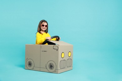 Photo of Little girl in sunglasses driving car made with cardboard on light blue background. Space for text