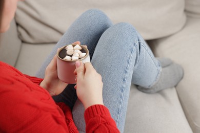 Photo of Woman holding mug of tasty hot chocolate with marshmallows on sofa, closeup