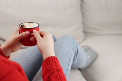 Photo of Woman holding mug of tasty hot chocolate with marshmallows on sofa, closeup. Space for text