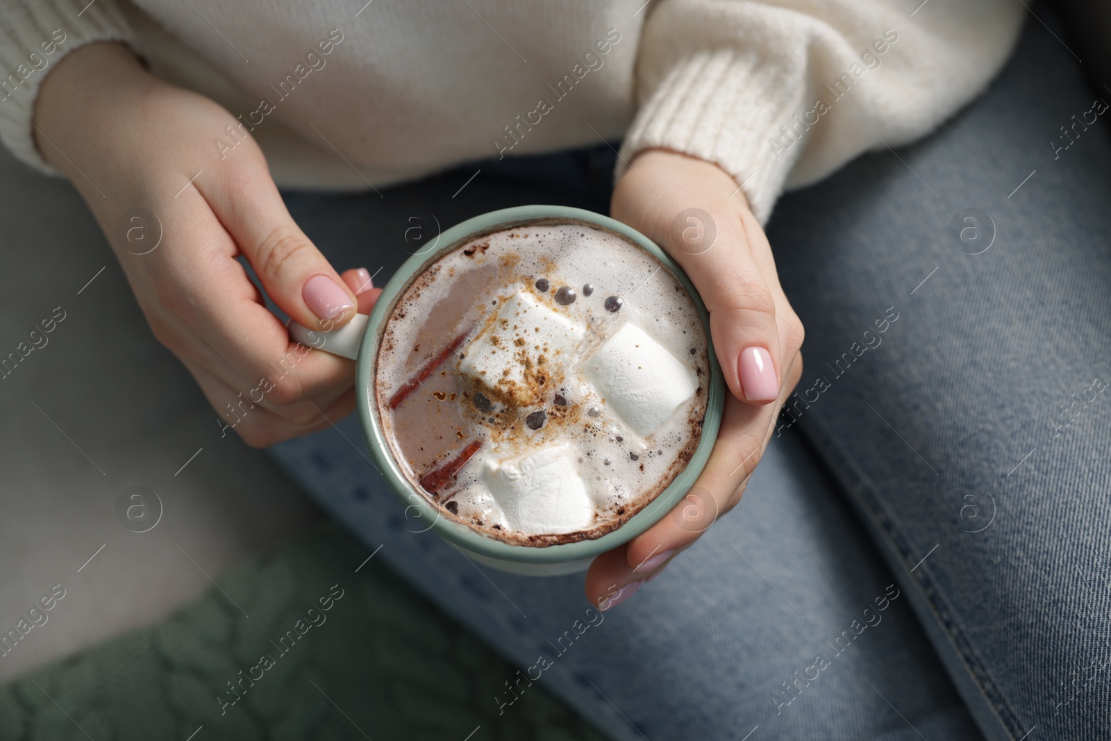 Photo of Woman holding mug of tasty hot chocolate with marshmallows and cinnamon on sofa, top view