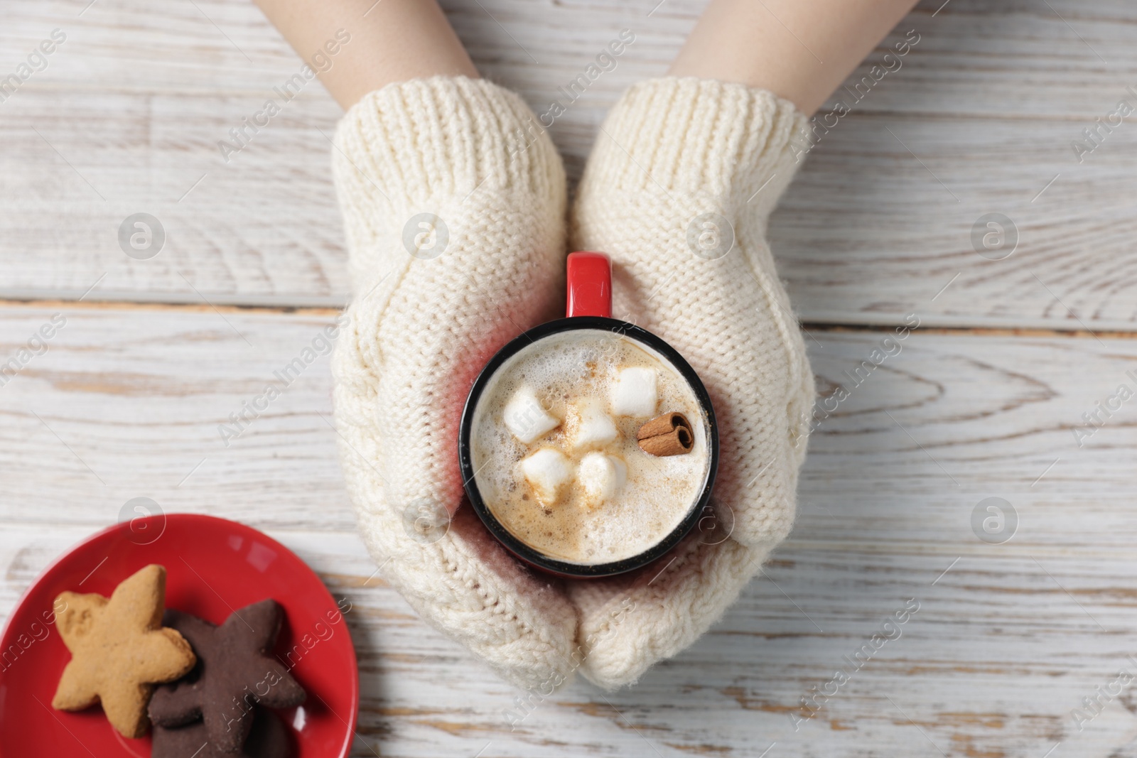 Photo of Woman having tasty hot chocolate with marshmallows, cinnamon and cookies at white wooden table, top view