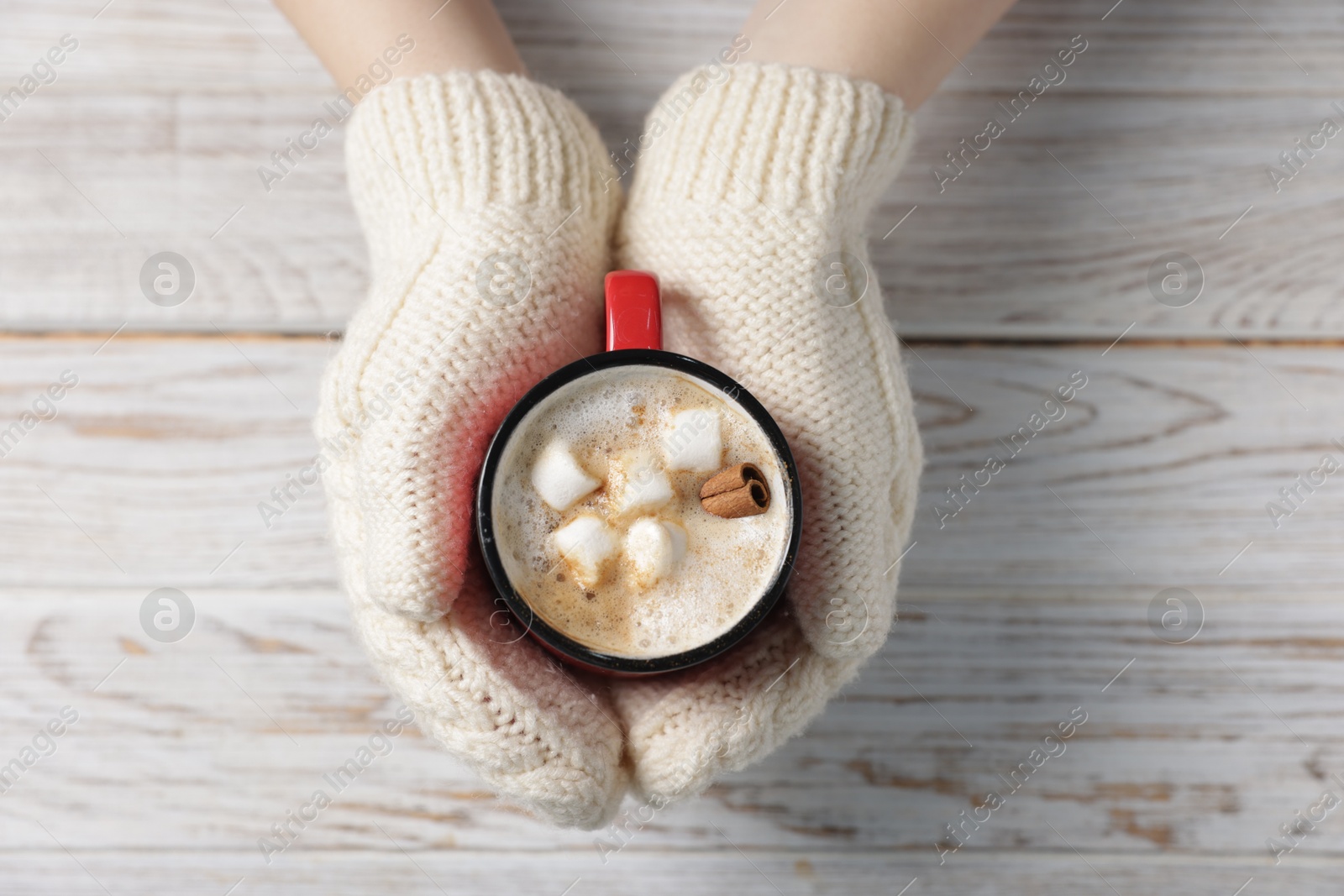 Photo of Woman holding mug of tasty hot chocolate with marshmallows and cinnamon at white wooden table, top view