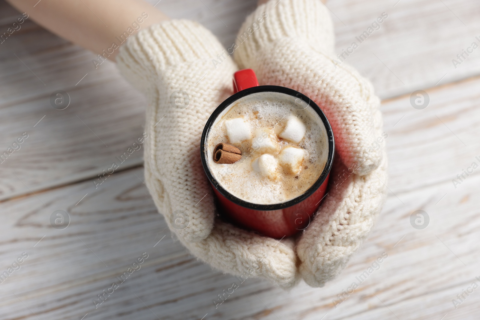 Photo of Woman holding mug of tasty hot chocolate with marshmallows and cinnamon at white wooden table, closeup