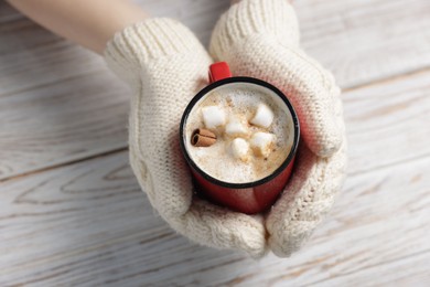 Photo of Woman holding mug of tasty hot chocolate with marshmallows and cinnamon at white wooden table, closeup