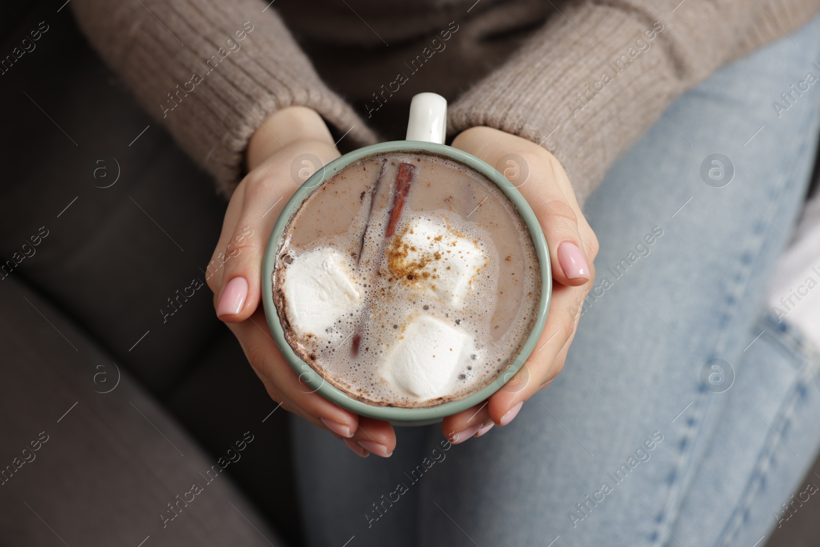 Photo of Woman holding mug of tasty hot chocolate with marshmallows and cinnamon indoors, top view