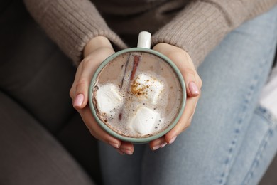 Photo of Woman holding mug of tasty hot chocolate with marshmallows and cinnamon indoors, top view