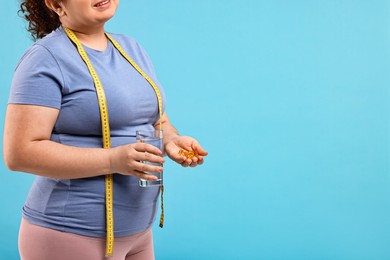 Photo of Happy plus size woman holding pile of weight loss supplements and glass with water on light blue background, closeup. Space for text